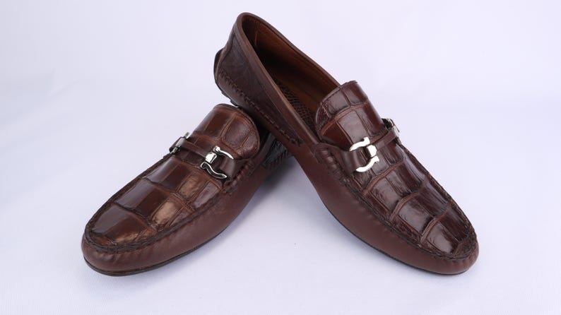 May include: A pair of brown leather loafers with a crocodile pattern. The shoes have silver-colored buckles and are displayed on a white background. These driving shoes are a classic style.