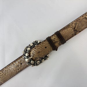 Op de afbeelding: Een riem met slangenpatroon en een zilverkleurige gesp. De riem heeft een bruine leren achterkant en een decoratieve gesp met een geconfronteerd ontwerp. De riem is bruin, beige en zilver van kleur.