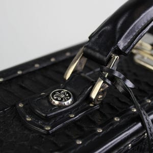 May include: Close-up of a black leather case with a handle and silver hardware. The case features a textured pattern, silver studs, and a decorative silver button with a fleur-de-lis design. A black strap is attached to the case.