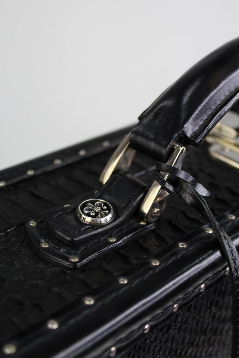 May include: Close-up of a black leather case with a handle and silver hardware. The case features a textured pattern, silver studs, and a decorative silver button with a fleur-de-lis design. A black strap is attached to the case.