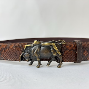 Può includere: Cintura in pelle marrone con fibbia a forma di toro color bronzo. La cintura ha un motivo a pelle di serpente. La fibbia presenta una scultura dettagliata di un toro con una figura che cavalca sulla sua schiena. Passante in pelle marrone abbinato.