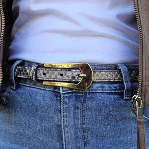 Può includere: Una cintura con motivo a pelle di serpente con una fibbia color oro è allacciata su un paio di jeans blu. La cintura è grigia e marrone con un motivo ripetuto. La fibbia dorata ha una forma curva.