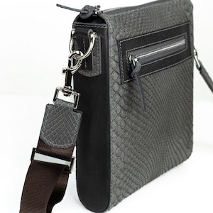 Puede incluir: Bolso bandolera de cuero gris con un patrón de piel de serpiente texturizado. El bolso presenta un panel negro, un bolsillo frontal con cremallera y una correa de hombro ajustable marrón. Los herrajes plateados incluyen un tirador de cremallera y fijaciones de correa.