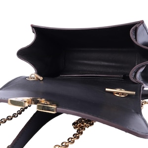 May include: Black leather handbag with a gold chain strap and clasp. The bag is open, revealing a dark interior. The edges are trimmed with brown leather. The bag has a unique, angular shape.