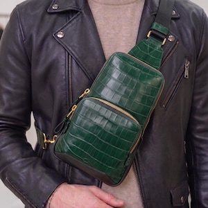 Puede incluir: Un bolso bandolera de color verde oscuro con un patrón texturizado de piel de cocodrilo, cremalleras de metal dorado y una correa de tela negra ajustable. El bolso tiene un compartimento principal y un bolsillo frontal, sobre una chaqueta de cuero.