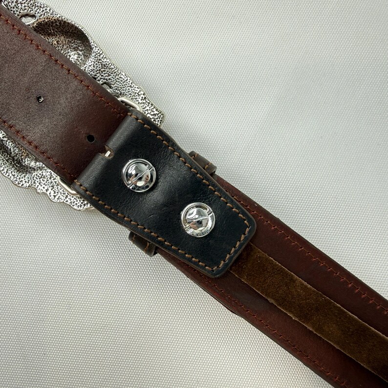 K&ouml;nnte beinhalten: Nahaufnahme eines braunen Lederg&uuml;rtels mit einer dekorativen Silberschnalle. Der G&uuml;rtel hat einen schwarzen Lederakzent mit zwei silbernen Nieten. Die N&auml;hte sind sichtbar und der G&uuml;rtel hat ein abgenutztes Aussehen.