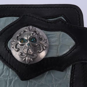 May include: Black leather wallet with a light blue, textured center panel. The wallet features a decorative silver skull emblem with turquoise accents and a black leather strap closure. The design is edgy and stylish.