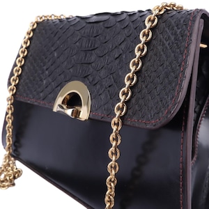 May include: Black handbag with a textured flap and a gold chain strap. The bag features a gold clasp and red stitching. The body of the bag is a glossy black, and the flap has a snakeskin pattern.