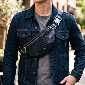 May include: A dark navy blue bum bag with a crocodile skin texture, featuring a metallic zip, a silver-toned skull accent on the strap, and a heavy-duty metal buckle closure. The bag is worn across the chest over a dark denim jacket and grey shirt.