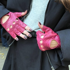 以下が含まれることがあります： 明るいピンク色の指なし手袋。ヘビ革のような質感で、指関節に3つの丸い切り抜きがあり、手のひらには大きな切り抜きがあります。黒いジャケットとグレーのセーターと合わせて着用されています。