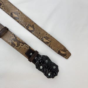 May include: A brown and tan snakeskin belt with a black buckle. The buckle has a textured design with black and clear stones. The belt is made of leather and has a classic design.
