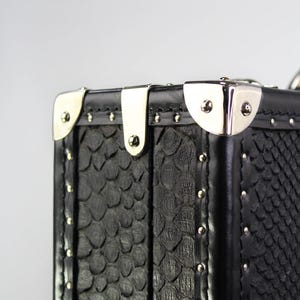 May include: Close-up of a black, textured trunk with silver-colored metal accents. The trunk features a scale-like pattern and is adorned with silver rivets and corner protectors. The design is classic and elegant.