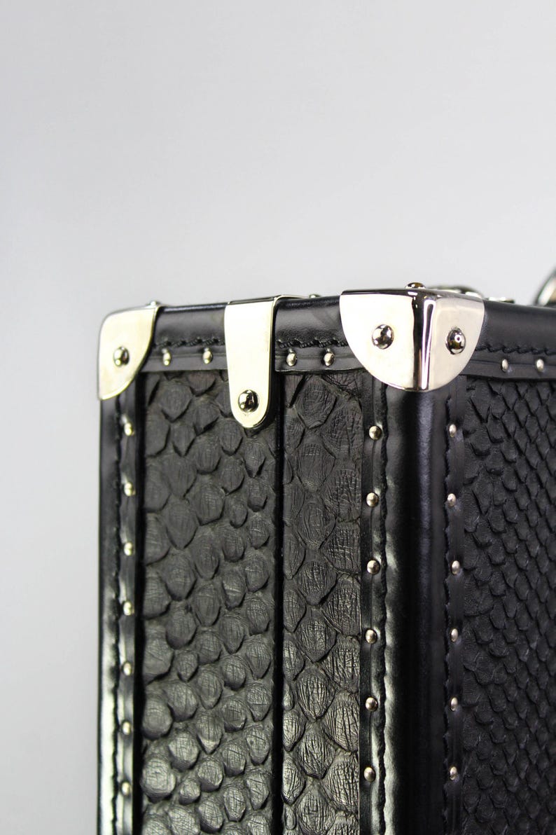 May include: Close-up of a black, textured trunk with silver-colored metal accents. The trunk features a scale-like pattern and is adorned with silver rivets and corner protectors. The design is classic and elegant.