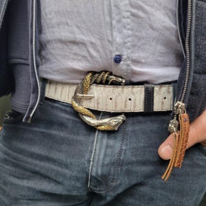 May include: A white leather belt with a snake-shaped buckle. The buckle is silver and gold, with the snake's head and tail forming the clasp. The belt is worn with blue jeans and a light-colored shirt.