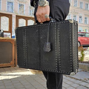 May include: A black briefcase with a snakeskin pattern, silver corner protectors, and studded trim. The briefcase has a leather handle and a matching tag. The case is rectangular and appears to be made of leather.