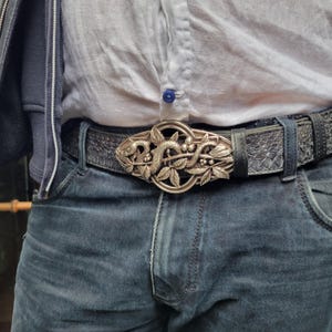 Könnte beinhalten: Silberfarbene Gürtelschnalle mit detailliertem Design einer Schlange, Blättern und Beeren. Die Schnalle ist an einem dunkelgrauen Gürtel mit Schlangenmuster befestigt, der mit einer blauen Jeans und einem weißen Hemd getragen wird.