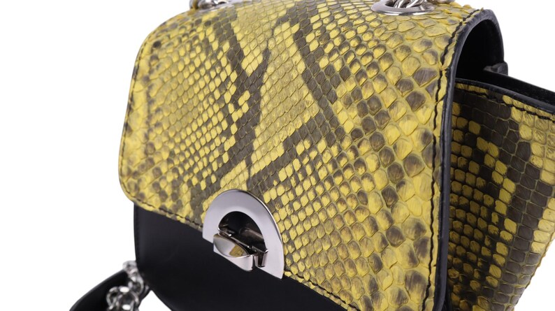 May include: A small, round handbag with a yellow and black snakeskin pattern on the top flap. The bottom portion of the bag is black, and it features a silver-colored clasp and chain strap. The bag is designed for carrying essentials.