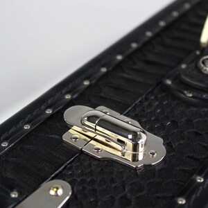 May include: Close-up of a black trunk with a textured surface and silver hardware. The trunk features a metal clasp and rivets along the edges. The design is reminiscent of vintage travel cases.