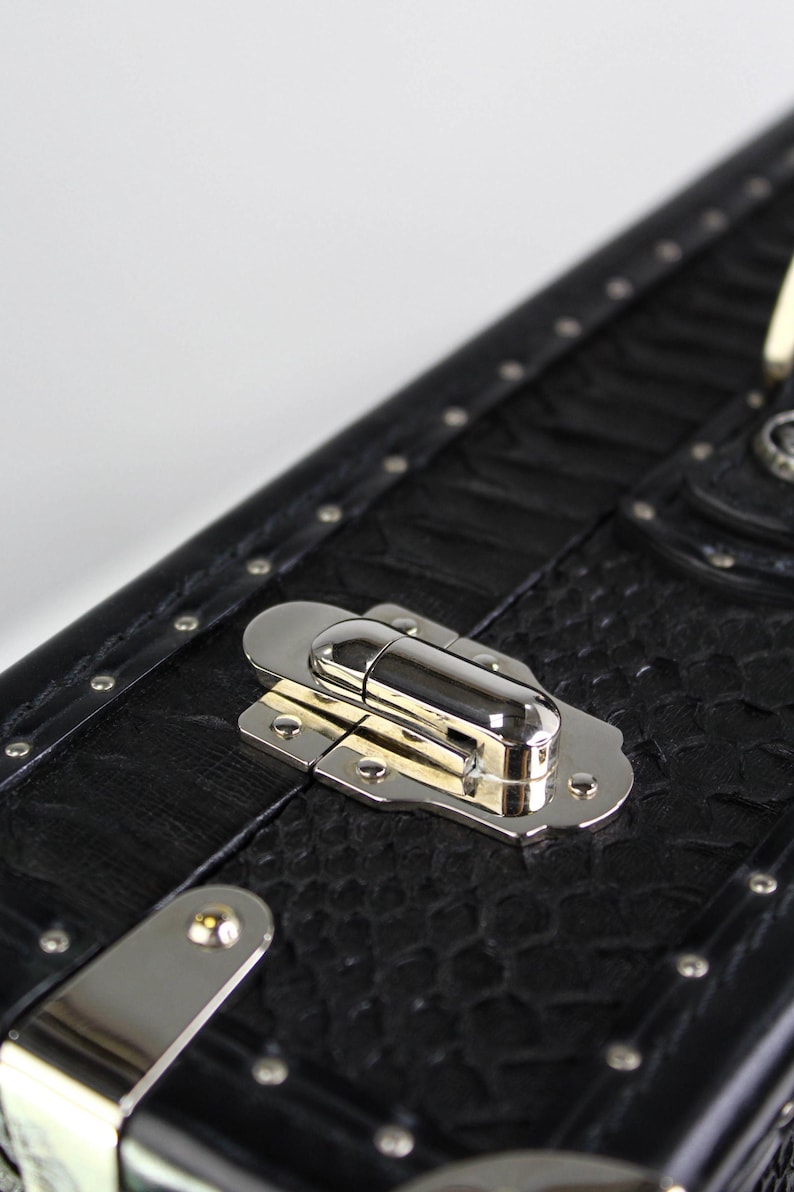 May include: Close-up of a black trunk with a textured surface and silver hardware. The trunk features a metal clasp and rivets along the edges. The design is reminiscent of vintage travel cases.