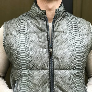 Könnte beinhalten: Eine silbergraue Weste mit Schlangenmuster. Die Weste hat einen hohen Kragen und einen durchgehenden Reißverschluss. Das Material scheint Leder oder ein lederähnliches Material zu sein. Die ärmellose Weste hat ein gestepptes Design.