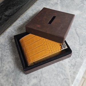 Pode incluir: Uma carteira amarelo mostarda com textura de pele de crocodilo é exibida dentro de uma caixa marrom com interior preto. A carteira tem uma peça decorativa prateada no canto. A tampa da caixa está ligeiramente aberta.