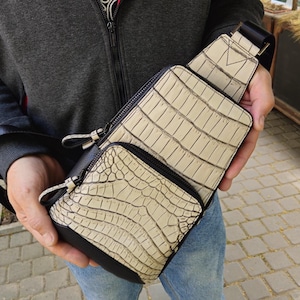 Op de afbeelding: Crèmekleurige crossbodytas met een krokodillenleerpatroon. De tas heeft een zwarte band en rits, en een kleinere voorzak. De tas wordt vastgehouden door een persoon die een grijze jas en een blauwe spijkerbroek draagt.