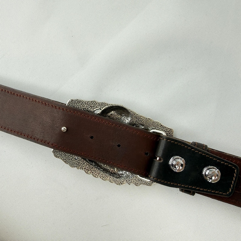 K&ouml;nnte beinhalten: Brauner Lederg&uuml;rtel mit einer gro&szlig;en, verzierten silberfarbenen Schnalle. Die Schnalle hat ein detailliertes, strukturiertes Design. Der G&uuml;rtel hat eine schwarze Lederschlaufe und silberfarbene Nieten. Der G&uuml;rtel wird vor wei&szlig;em Hintergrund gezeigt.