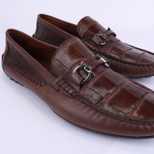 May include: A pair of brown leather loafers with a crocodile-like texture on the vamp. The shoes have silver-colored metal accents and a stitched design on the heel. The loafers are displayed on a white surface.