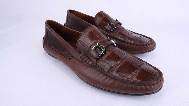 May include: A pair of brown leather loafers with a crocodile-like texture on the vamp. The shoes have silver-colored metal accents and a stitched design on the heel. The loafers are displayed on a white surface.