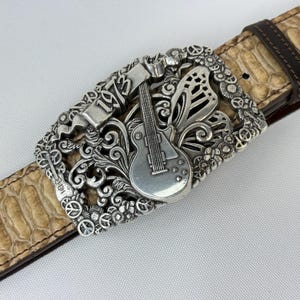 K&ouml;nnte beinhalten: Silberfarbene G&uuml;rtelschnalle mit Gitarre, dem Jahr "1973" und floralen und Peace-Zeichen-Akzenten. Die Schnalle ist an einem hellbraunen, strukturierten G&uuml;rtel mit dunkelbraunem Lederfutter befestigt.
