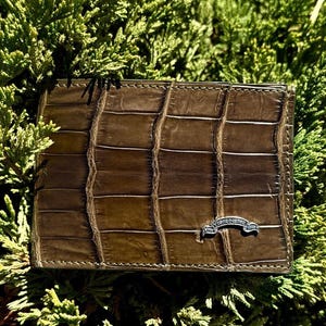 Puede incluir: Una cartera rectangular de color marrón con un patrón de textura de piel de caimán. La cartera tiene costuras visibles a lo largo de los bordes y un pequeño emblema metálico plateado curvado en la parte frontal con el texto "VTOOR DESIGN SINCE 1998".