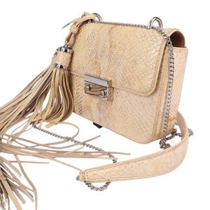 Peut inclure: Sac bandoulière beige avec un motif peau de serpent. Il est doté d'une fermeture à rabat avec un fermoir argenté, d'une bandoulière chaîne argentée et d'une longue gland. Le sac est de couleur neutre et a un aspect texturé.