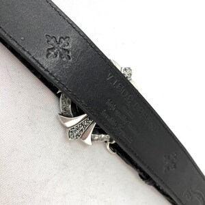 May include: Black leather belt with a silver buckle featuring a cross design and rhinestones. The belt has embossed floral patterns and the text "VIGOR DESIGN" and "high quality leather" on the inside.