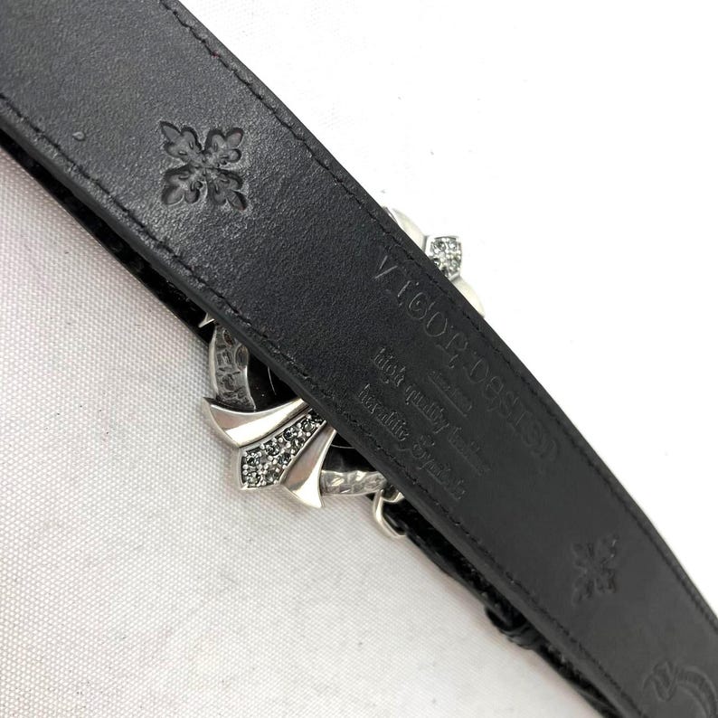 May include: Black leather belt with a silver buckle featuring a cross design and rhinestones. The belt has embossed floral patterns and the text "VIGOR DESIGN" and "high quality leather" on the inside.