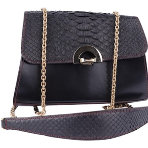 May include: Black handbag with a textured flap and a gold chain strap. The bag has a sleek, rectangular design with a contrasting smooth leather body. The strap and flap have a reptile-like pattern. A gold-tone clasp secures the bag.