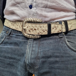 Könnte beinhalten: Ein Gürtel mit Schlangenmuster und silberfarbener Schnalle wird über einer blauen Jeans getragen. Der Gürtel hat ein cremefarbenes und braunes Muster mit schwarzen Lederakzenten. Die Schnalle hat ein dekoratives, geriffeltes Design.