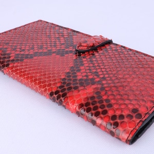 May include: A red and black snakeskin-patterned wallet with a snap closure. The wallet is rectangular and has a black border. The snakeskin pattern is a mix of red and black scales.