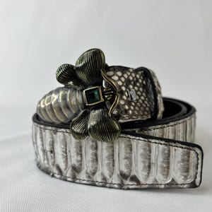 Puede incluir: Un cinturón con estampado de piel de serpiente con una hebilla decorativa. La hebilla es una abeja plateada con una gema cuadrada verde en el centro. El cinturón es blanco y gris con interior negro.