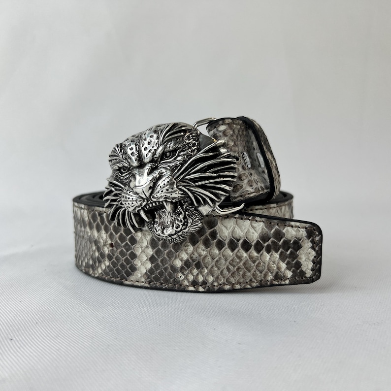 Pode incluir: Um cinto com padr&atilde;o de pele de cobra em tons de cinza e branco. O cinto apresenta uma fivela prateada em forma de cabe&ccedil;a de tigre rugindo. O cinto est&aacute; enrolado sobre uma superf&iacute;cie branca.