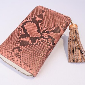 May include: A pink and brown snakeskin-patterned wallet with a zipper closure and a tassel. The wallet is rectangular and has a light-colored zipper. The tassel is made of leather and has a gold-colored cap.