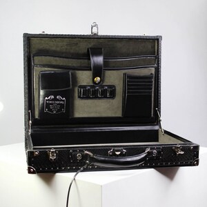 May include: Open black leather briefcase with a textured exterior. The interior features multiple pockets, a card holder, and a flap closure. The briefcase is on a white surface.