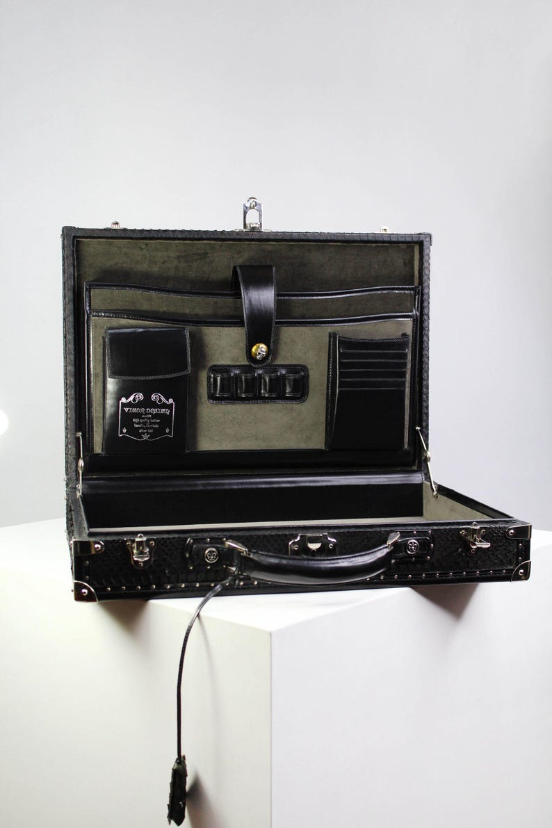 May include: Open black leather briefcase with a textured exterior. The interior features multiple pockets, a card holder, and a flap closure. The briefcase is on a white surface.