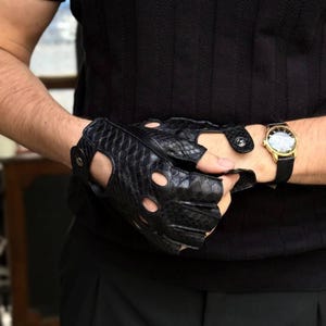 Puede incluir: Guantes sin dedos de cuero negro con un patrón de piel de serpiente texturizado. Los guantes tienen detalles recortados y un cierre a presión. También se ve un reloj dorado con una correa negra.