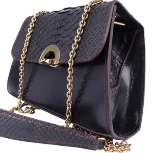 May include: Black leather shoulder bag with a textured flap and gold-tone chain strap. The bag has a rectangular shape with a flap closure and a gold-tone clasp. The strap is a combination of leather and chain.