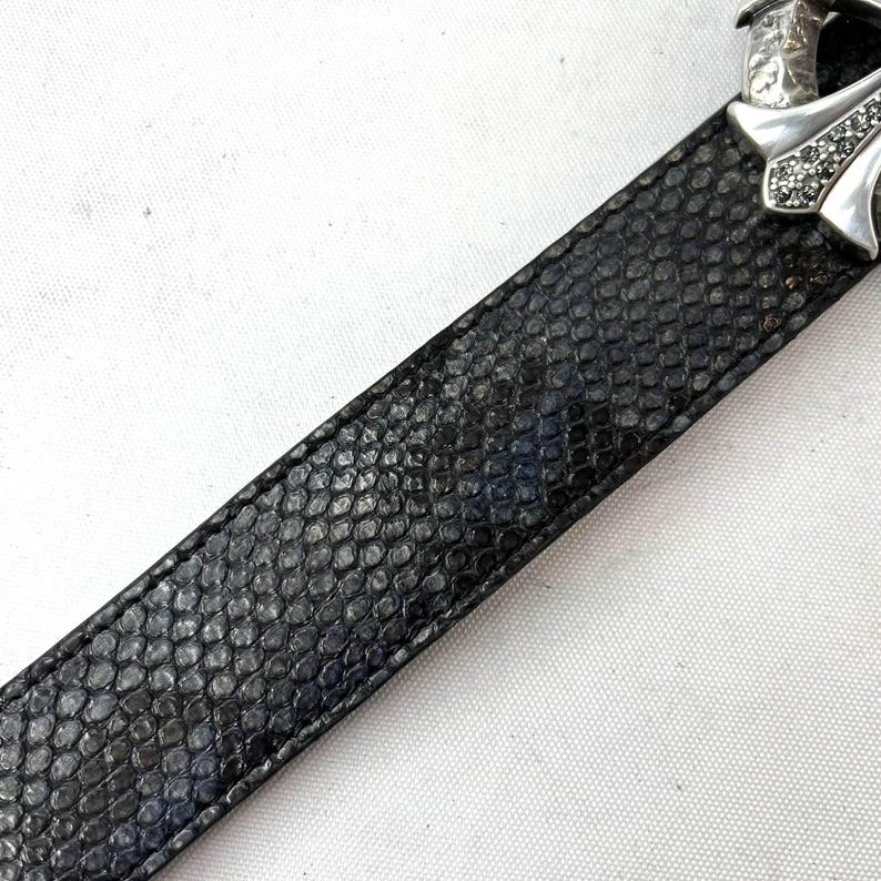 May include: A black leather belt with a textured, scale-like pattern. The belt has a silver-coloured buckle with a cross design, embellished with small, sparkling stones. The belt is angled across a white surface.