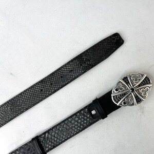 K&ouml;nnte beinhalten: Ein schwarzer G&uuml;rtel mit einem strukturierten Schlangenmuster. Der G&uuml;rtel hat eine silberfarbene Schnalle mit einem dekorativen Design und schwarzen Akzenten. Der G&uuml;rtel ist aus Leder und hat einen klassischen Stil.