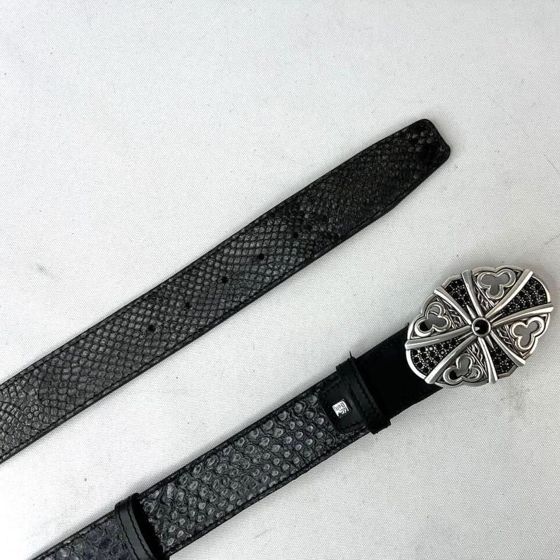 K&ouml;nnte beinhalten: Ein schwarzer G&uuml;rtel mit einem strukturierten Schlangenmuster. Der G&uuml;rtel hat eine silberfarbene Schnalle mit einem dekorativen Design und schwarzen Akzenten. Der G&uuml;rtel ist aus Leder und hat einen klassischen Stil.