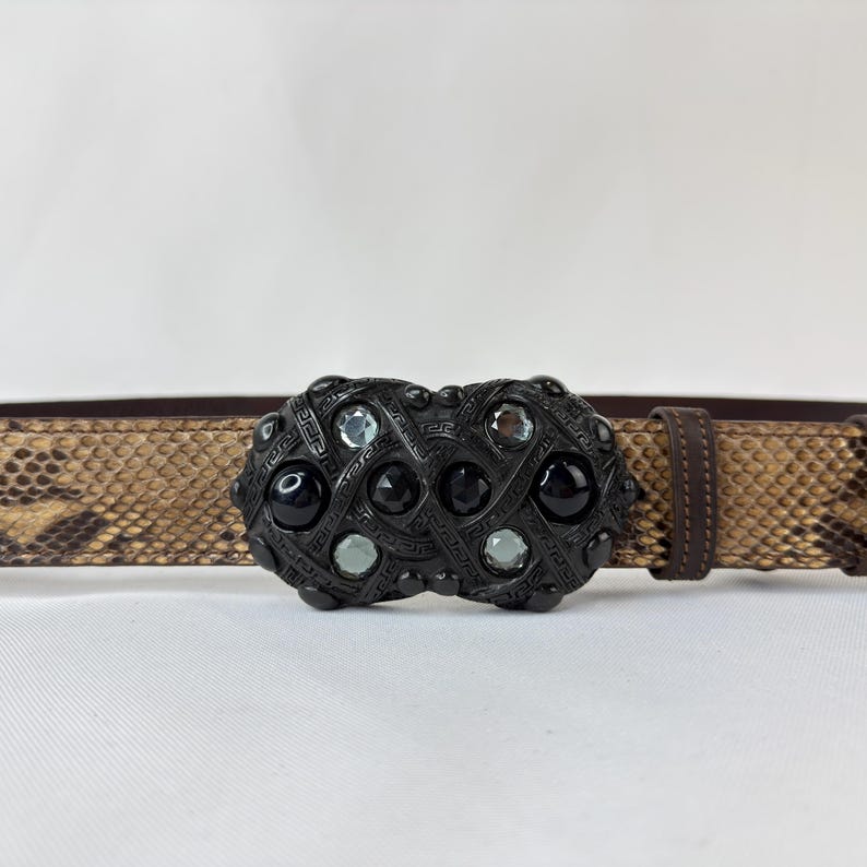 May include: A brown belt with a snakeskin pattern and a decorative black buckle. The buckle features black and clear gemstones. The belt is made of leather and has a classic design.
