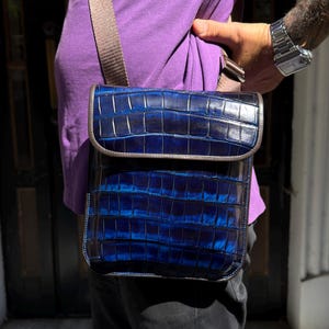 May include: A rectangular crossbody bag featuring a deep blue, glossy crocodile-embossed texture with a flap closure. The bag includes a wide, neutral-toned fabric shoulder strap that connects to the top edges, providing a structured appearance.