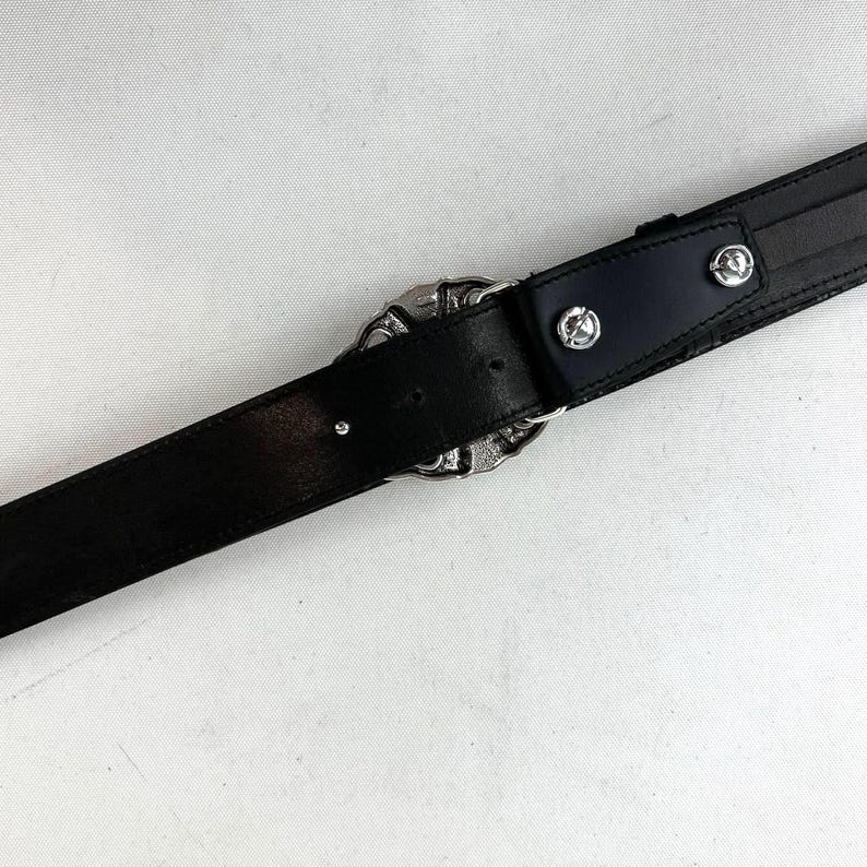 K&ouml;nnte beinhalten: Schwarzer Lederg&uuml;rtel mit einer silberfarbenen, verzierten Schnalle. Der G&uuml;rtel hat ein klassisches Design mit einer detaillierten Schnalle und silbernen Nieten. Ein vielseitiges Accessoire.